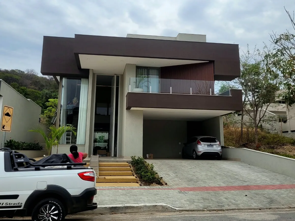 Fachada de uma casa de dois pavimentos pronta: volume de cima marrom escuro sobre a garagem, painel de madeira ao lado do guarda-corpo de vidro, um carro prata na garagem e uma picape branca na frente