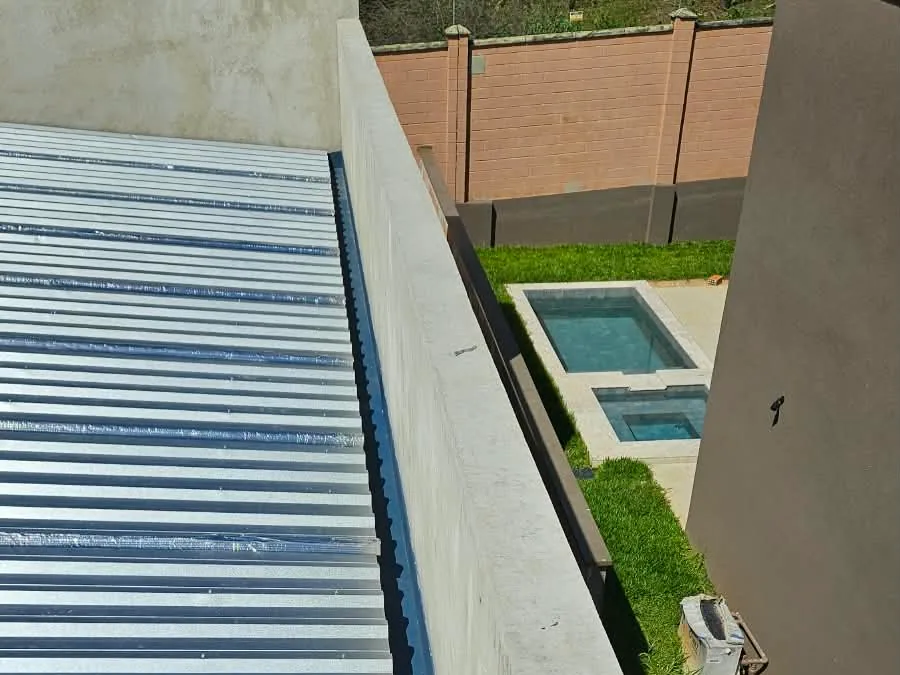 Telhado de telha metálica sobre laje, ao lado de uma platibanda de concreto, com piscina e jardim ao fundo