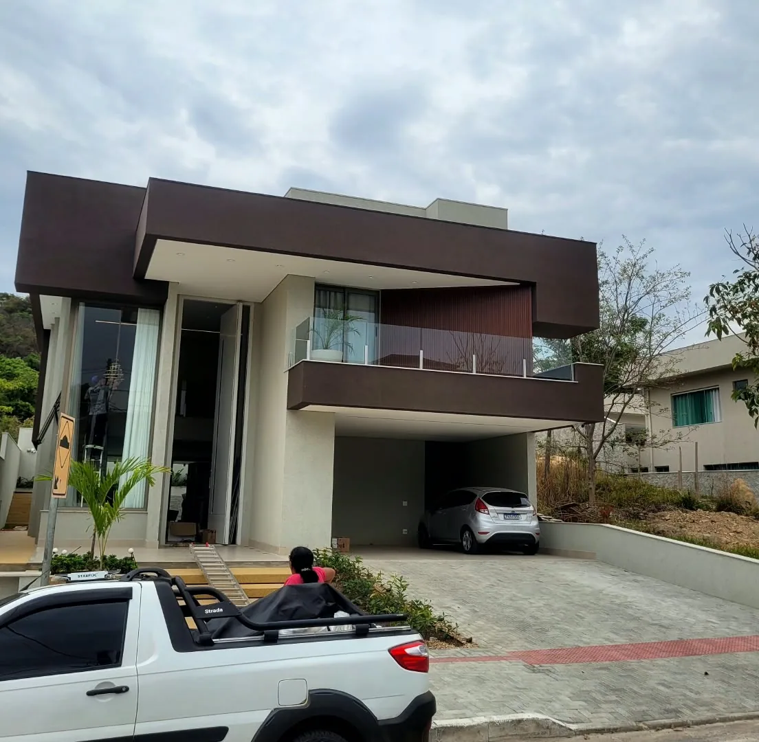 Casa de dois pavimentos com o volume de cima revestido em marrom escuro sobre a garagem, fachada clara com vidro e guarda-corpo de vidro, e uma picape branca na frente