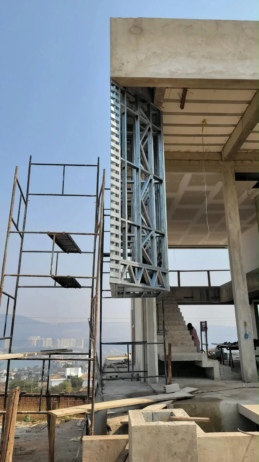 Estrutura de perfis de aço do light steel frame em balanço sob a laje de concreto de uma casa em obra, com andaime ao lado e a cidade ao fundo sob céu azul