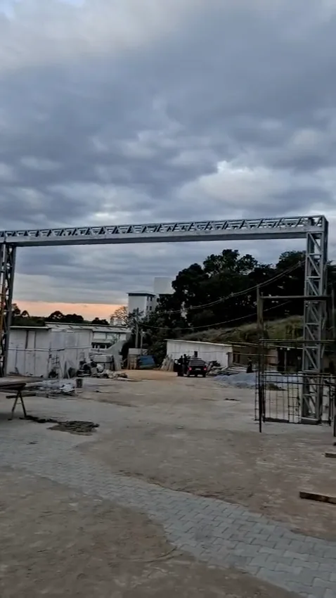 Quadro do vídeo: pórtico de treliças de aço montado num canteiro de obra ao entardecer, com céu nublado