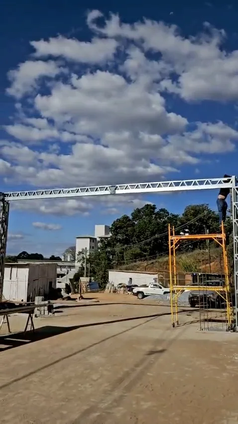 Quadro do vídeo: viga em treliça de aço no alto de uma torre, com andaime amarelo e um operário no topo, sob céu azul